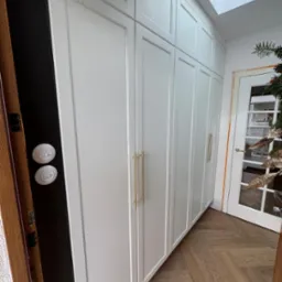 Wysokie, białe szafy na wymiar z delikatnymi, złotymi uchwytami, zamontowane w wąskim przedpokoju z parkietem ułożonym w jodełkę, widoczne drzwi z szybami i fragment dekoracji świątecznej.