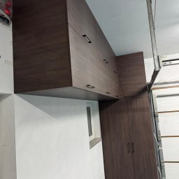 WOJCIECH BIESIADA - Ciemnobrązowe szafy na wymiar z podłużnymi uchwytami, zamontowane pod sufitem w garażu, widoczne okno i brama garażowa.