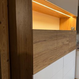 WOJCIECH BIESIADA - Fragment szafy z drewnianymi frontami i białymi szufladami, oświetlenie LED pod górną szafką.