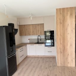 WOJCIECH BIESIADA - Nowoczesna kuchnia z jasnymi szafkami dolnymi, drewnopodobnymi szafkami górnymi, czarnym zlewem i lodówką, oraz niedokończonym oświetleniem sufitowym.