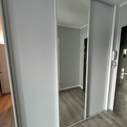 WOJCIECH BIESIADA - Nowoczesna szafa przesuwna z lustrzanymi drzwiami, szare panele podłogowe, widok na wnętrze pokoju przez lustro.