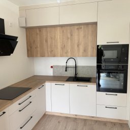 WOJCIECH BIESIADA - Nowoczesna kuchnia z białymi szafkami dolnymi, drewnopodobnymi szafkami górnymi, czarnym zlewem i płytą indukcyjną, oraz zabudowanym piekarnikiem i mikrofalówką marki Beko.