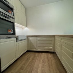 WOJCIECH BIESIADA - Jasna kuchnia z drewnianą podłogą i zabudową meblową w odcieniach beżu, widoczne dwa piekarniki Whirlpool z naklejkami promocyjnymi.