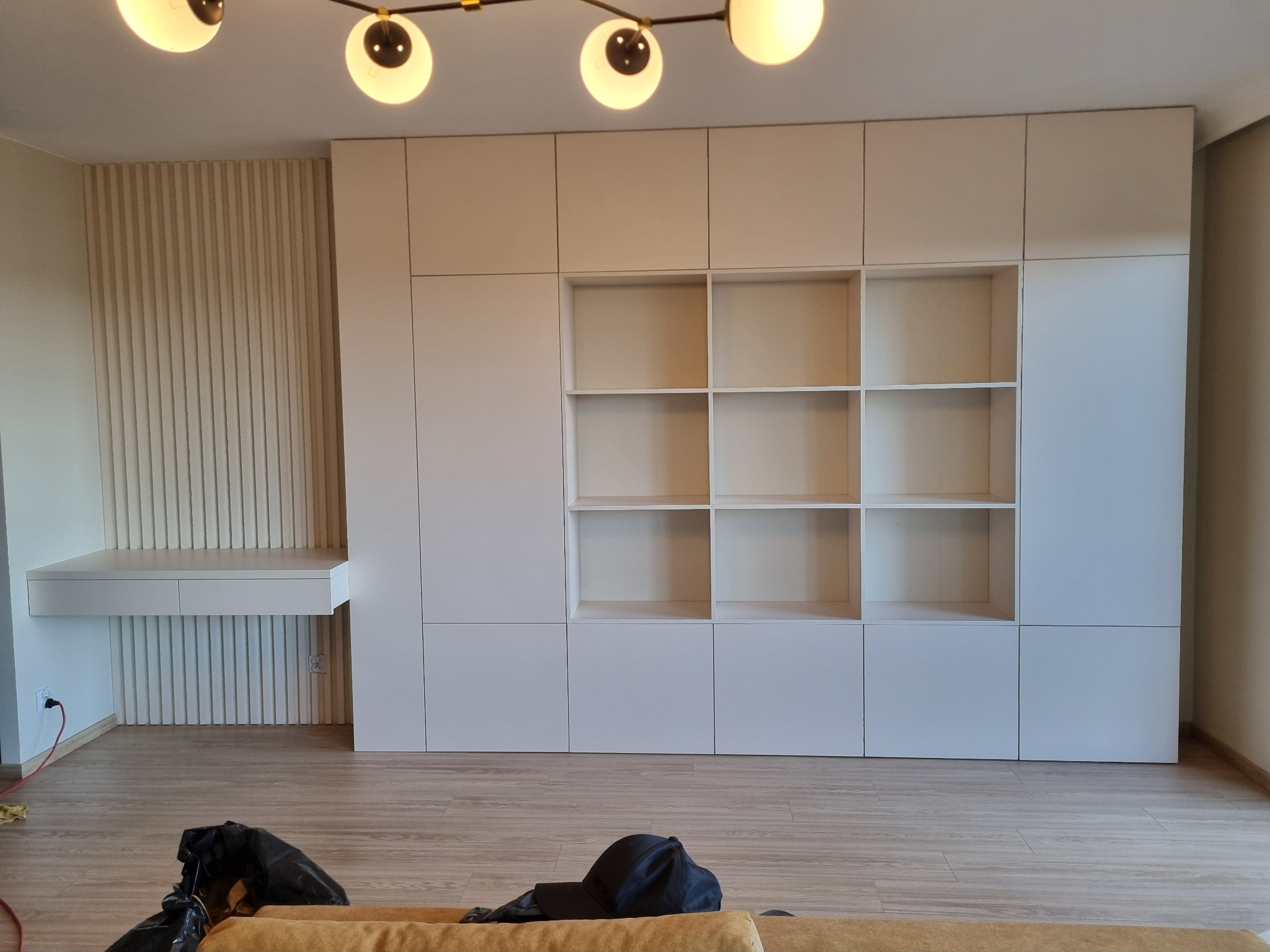 Biała zabudowa meblowa z szafkami i półkami, zintegrowana z biurkiem i dekoracyjnymi panelami ściennymi w pionowe prążki, oświetlona przez designerską lampę sufitową z kulistymi kloszami.