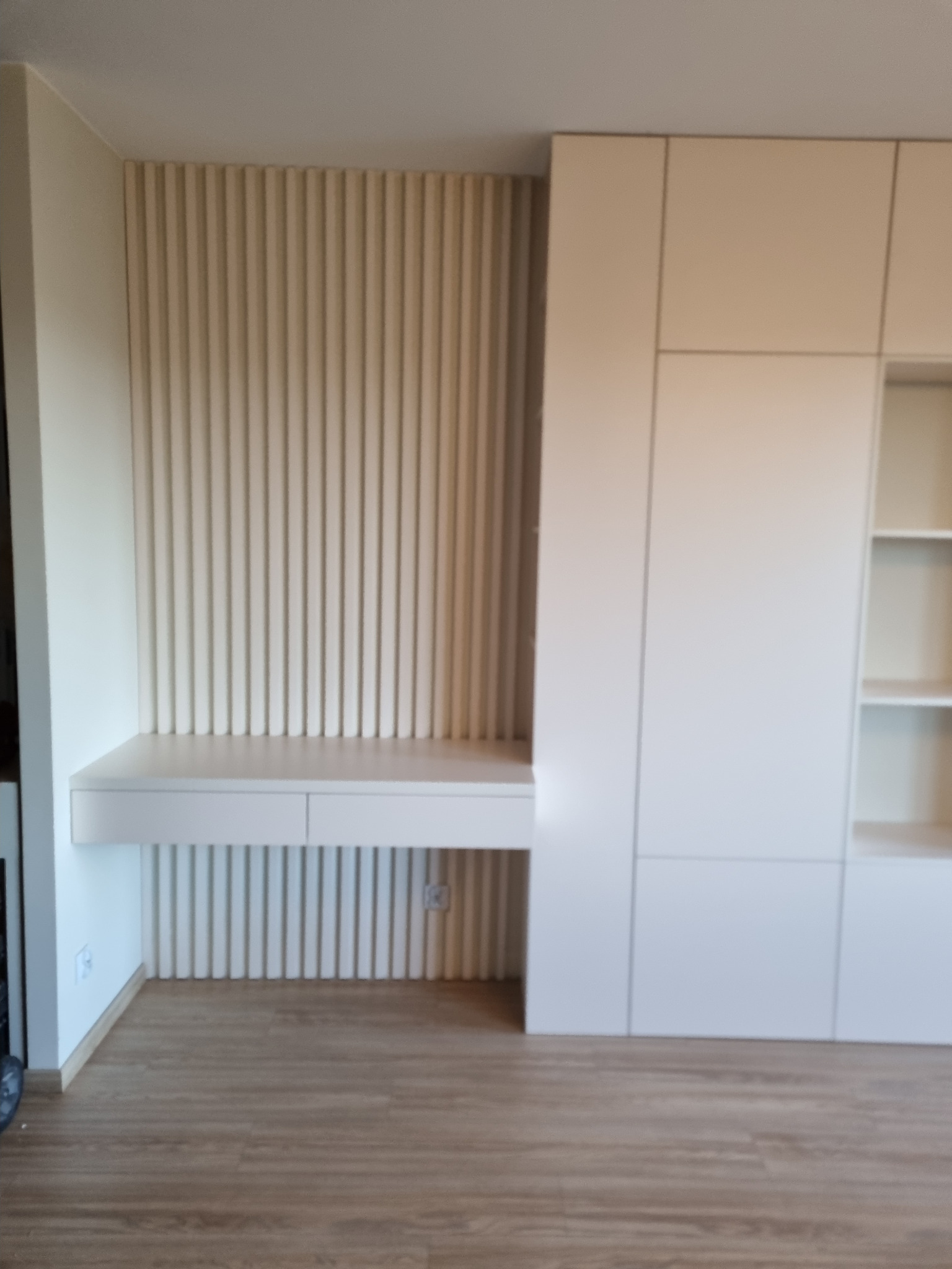 Biała szafa wnękowa z biurkiem w minimalistycznym stylu, z ryflowanym panelem ściennym w tle i drewnianą podłogą.