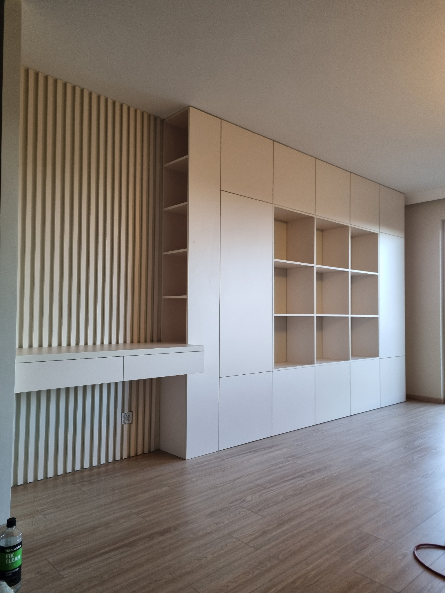 Biała szafa wnękowa z biurkiem i dekoracyjną ścianą z pionowych listew, widoczna butelka środka czyszczącego na podłodze.