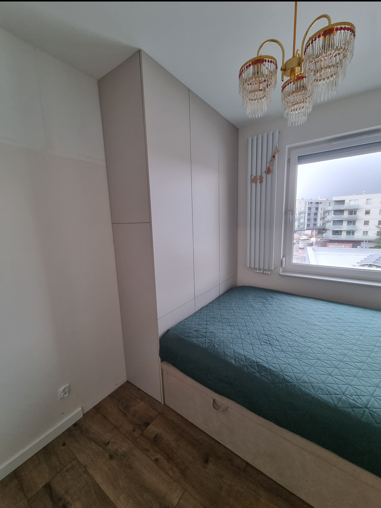 Wnęka sypialniana z zabudową szafy w kolorze beżowym, łóżko z szufladami i zielonym pikowanym materacem, widok na budynki mieszkalne za oknem, ozdobny żyrandol.