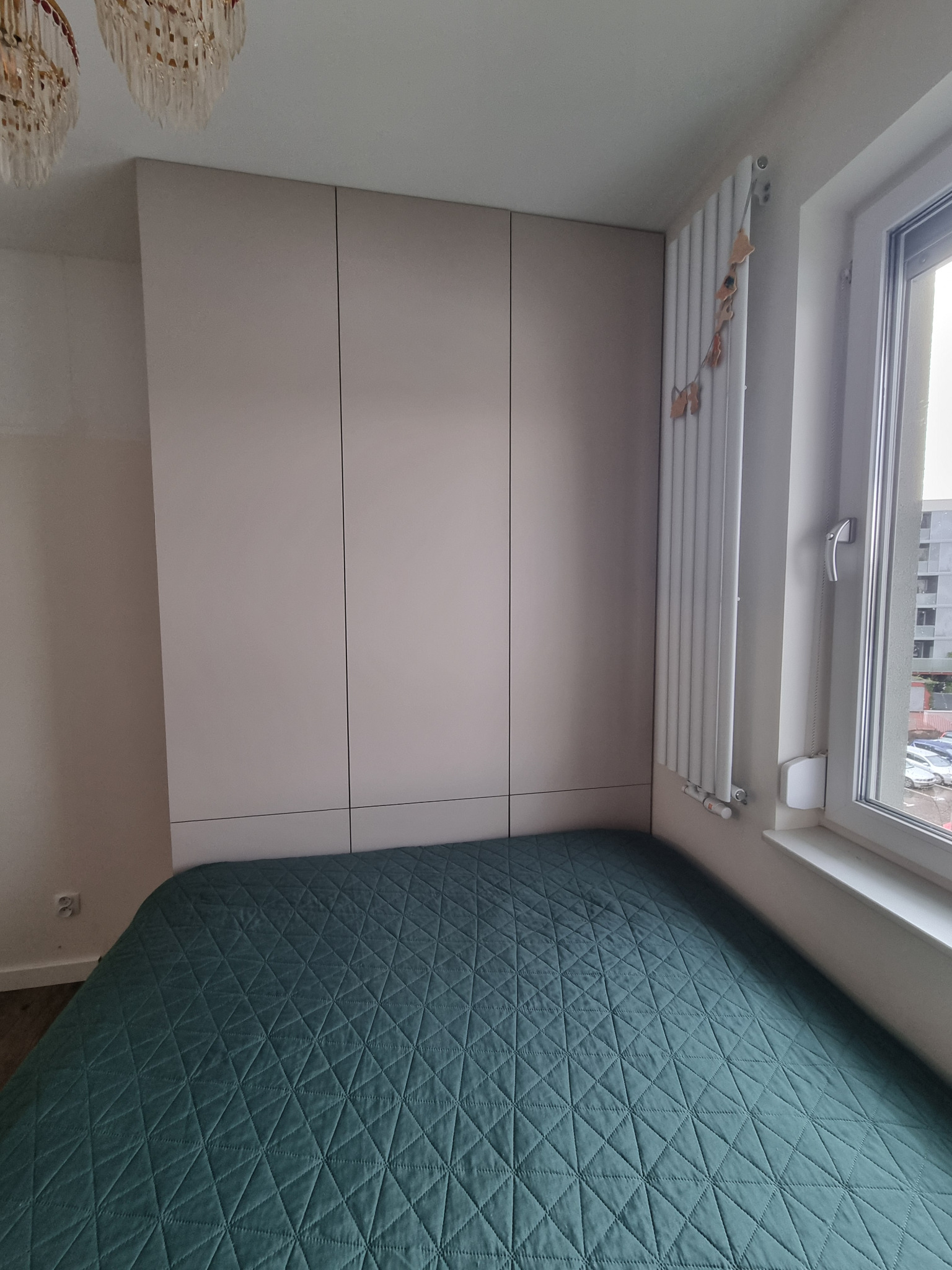 Szafa wnękowa z trzema drzwiami w kolorze beżowym, dopasowana do szerokości łóżka z zieloną narzutą w geometryczny wzór, widok na okno z grzejnikiem z dekoracją.