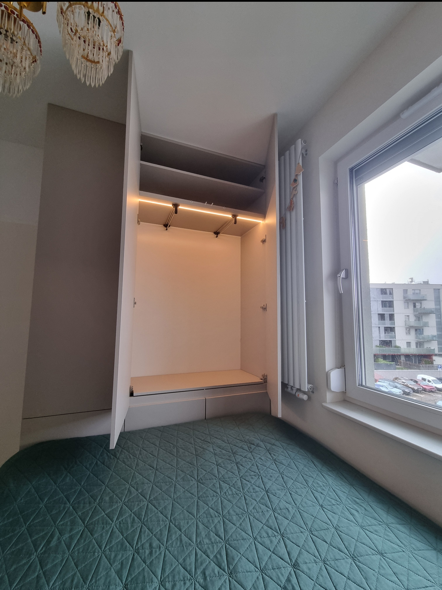 Wnętrze szafy wnękowej z oświetleniem LED, widok z dołu na puste półki i drążek, obok okno z widokiem na blok mieszkalny, zielona narzuta na łóżko na pierwszym planie.