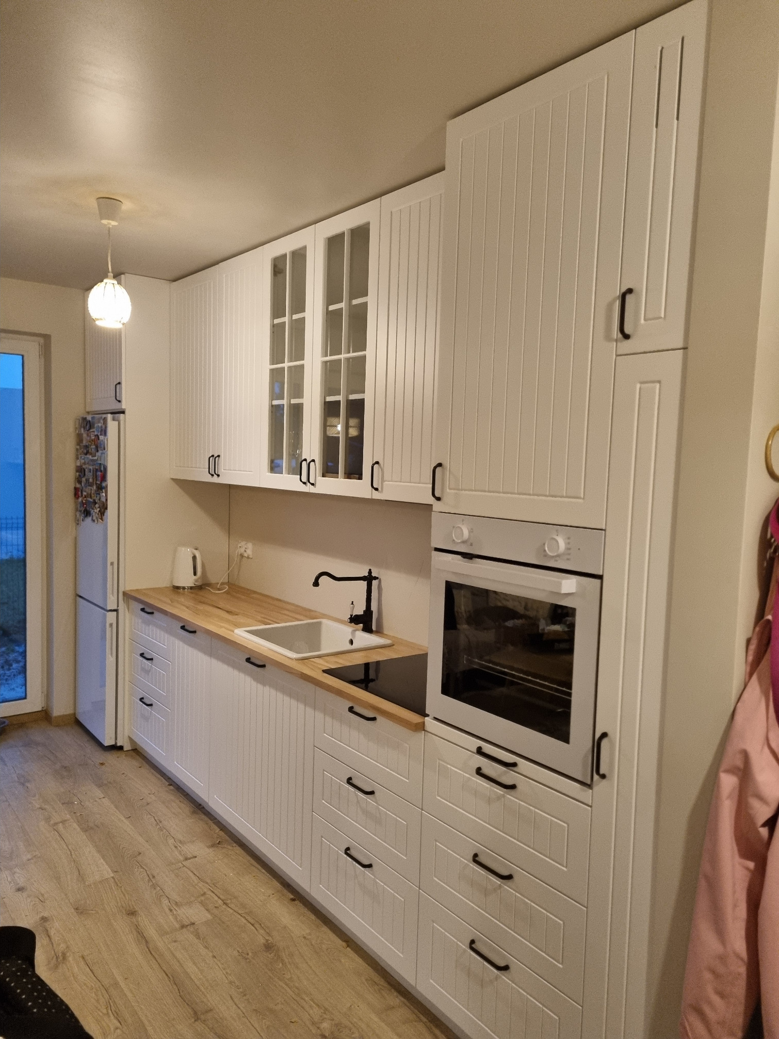 Biała, nowoczesna kuchnia z drewnianym blatem, zabudowaną piekarnikiem i szafkami z przeszklonymi frontami, oświetlona wiszącą lampą.