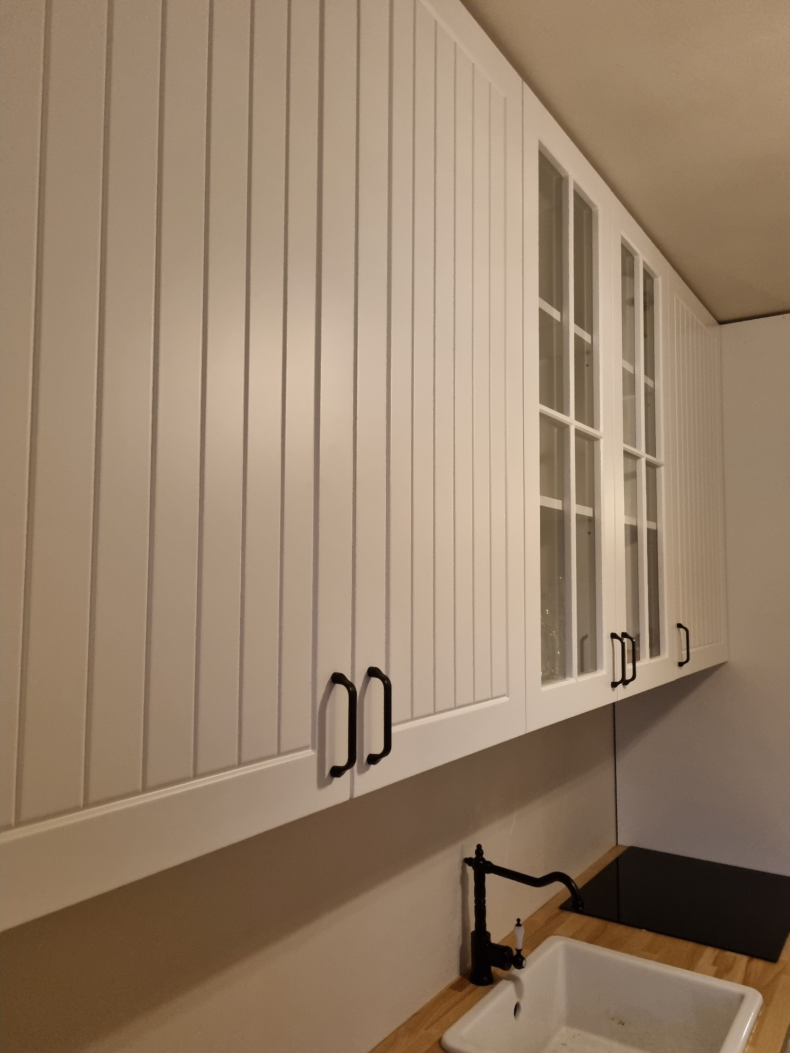 Białe szafki kuchenne wiszące z frezowanymi frontami i czarnymi uchwytami, część szafek z przeszklonymi drzwiczkami, widoczny zlew i czarna płyta grzewcza.