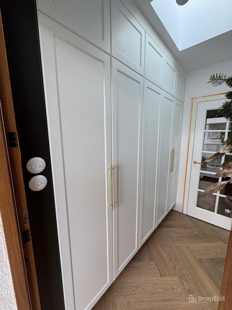 Wysokie, białe szafy na wymiar z delikatnymi, złotymi uchwytami, zamontowane w wąskim przedpokoju z parkietem ułożonym w jodełkę, widoczne drzwi z szybami i fragment dekoracji świątecznej.