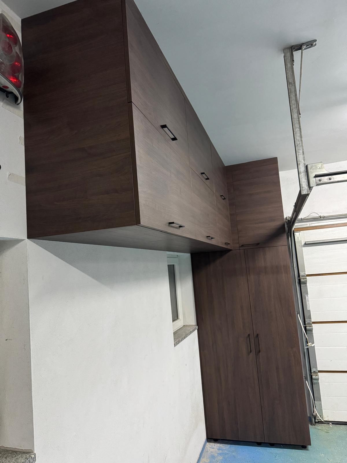Ciemnobrązowe szafy na wymiar z podłużnymi uchwytami, zamontowane pod sufitem w garażu, widoczne okno i brama garażowa.