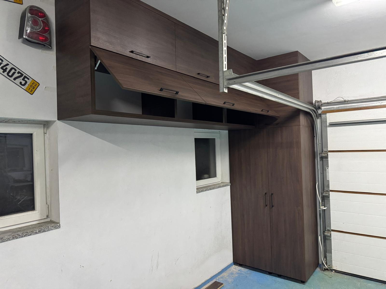 Szafki z ciemnego drewna zamontowane w garażu, widoczny fragment bramy garażowej i okno.