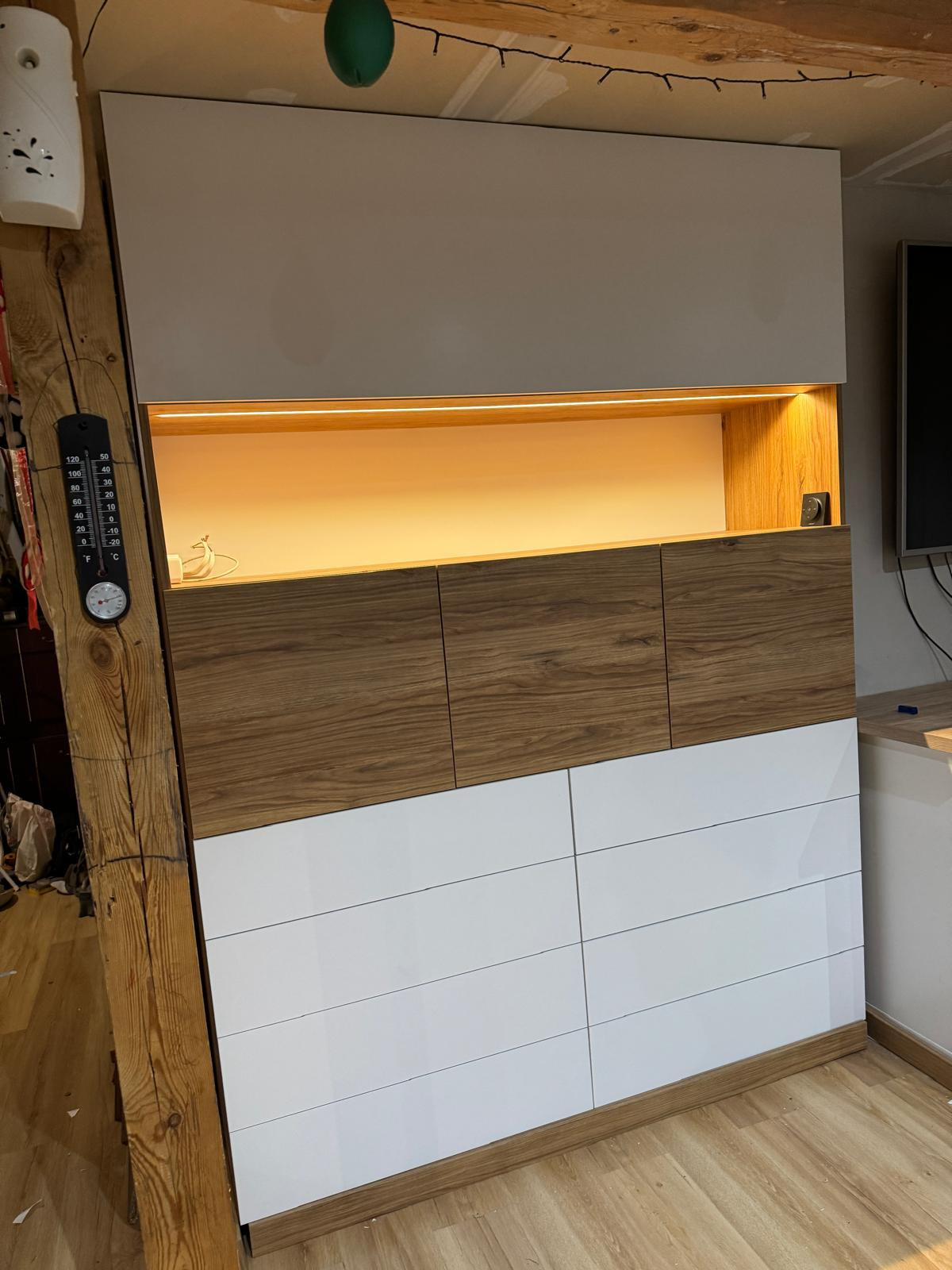 Biało-drewniana szafa z oświetleniem LED, wbudowana w nierówną ścianę z drewnianą belką i termometrem na niej.