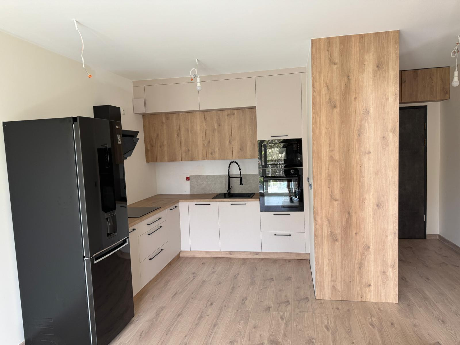 Nowoczesna kuchnia z jasnymi szafkami dolnymi, drewnopodobnymi szafkami górnymi, czarnym zlewem i lodówką, oraz niedokończonym oświetleniem sufitowym.