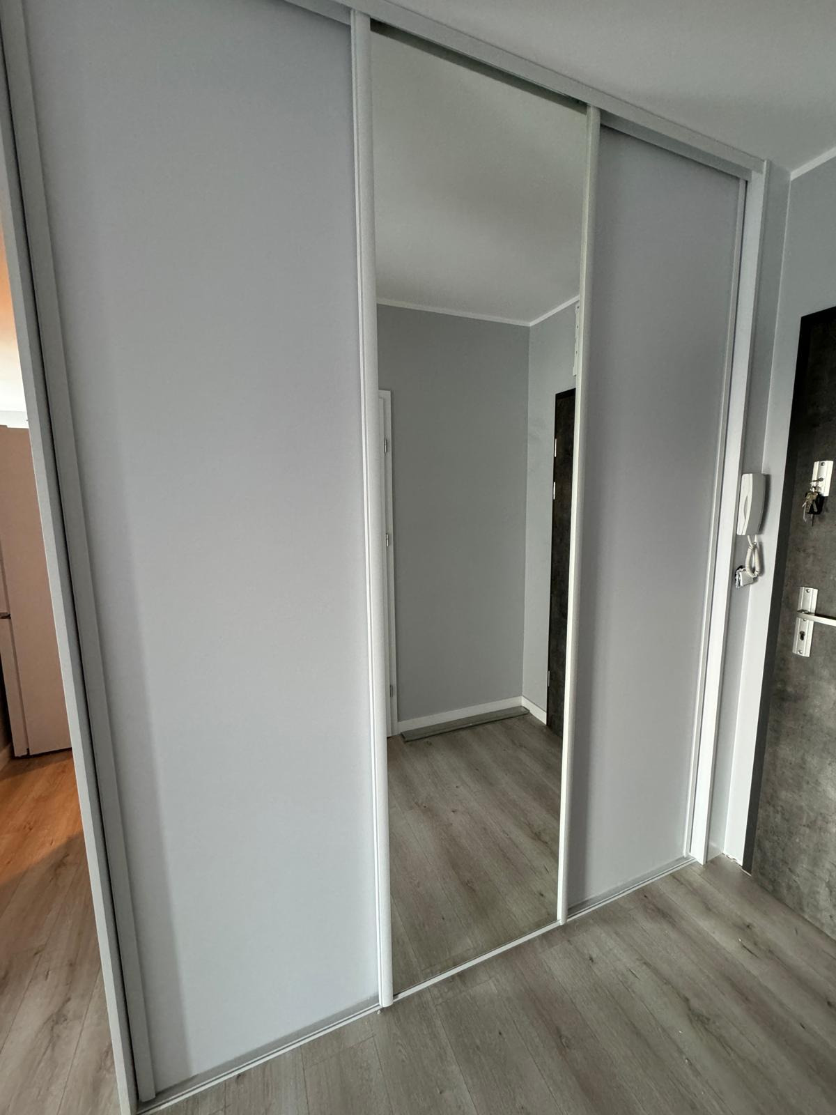 Nowoczesna szafa przesuwna z lustrzanymi drzwiami, szare panele podłogowe, widok na wnętrze pokoju przez lustro.