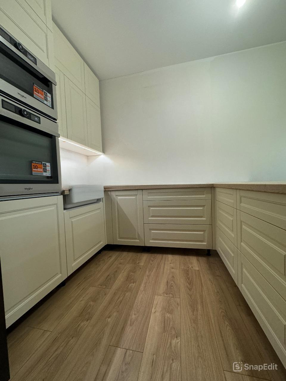 Jasna kuchnia z drewnianą podłogą i zabudową meblową w odcieniach beżu, widoczne dwa piekarniki Whirlpool z naklejkami promocyjnymi.