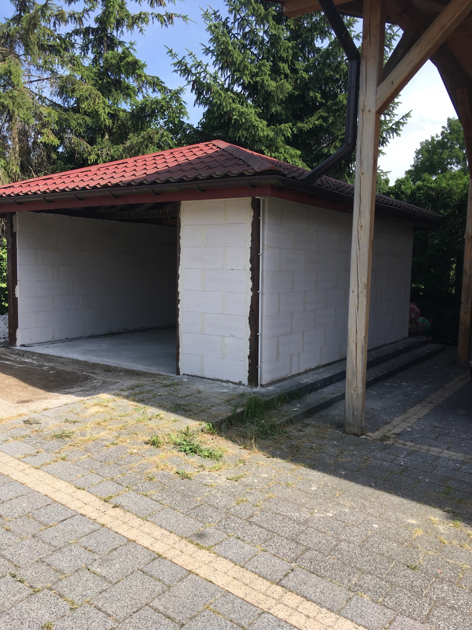 Surowy budynek gospodarczy z czerwonym dachem, zbudowany z białych bloczków, widoczny z zewnątrz na tle drzew.