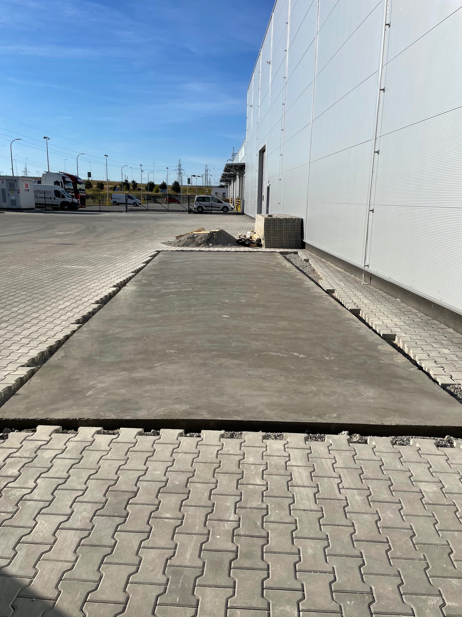 Świeżo wylana, prostokątna płyta betonowa na zewnątrz budynku przemysłowego, otoczona kostką brukową, z widoczną stertą piasku i materiałów budowlanych w tle.