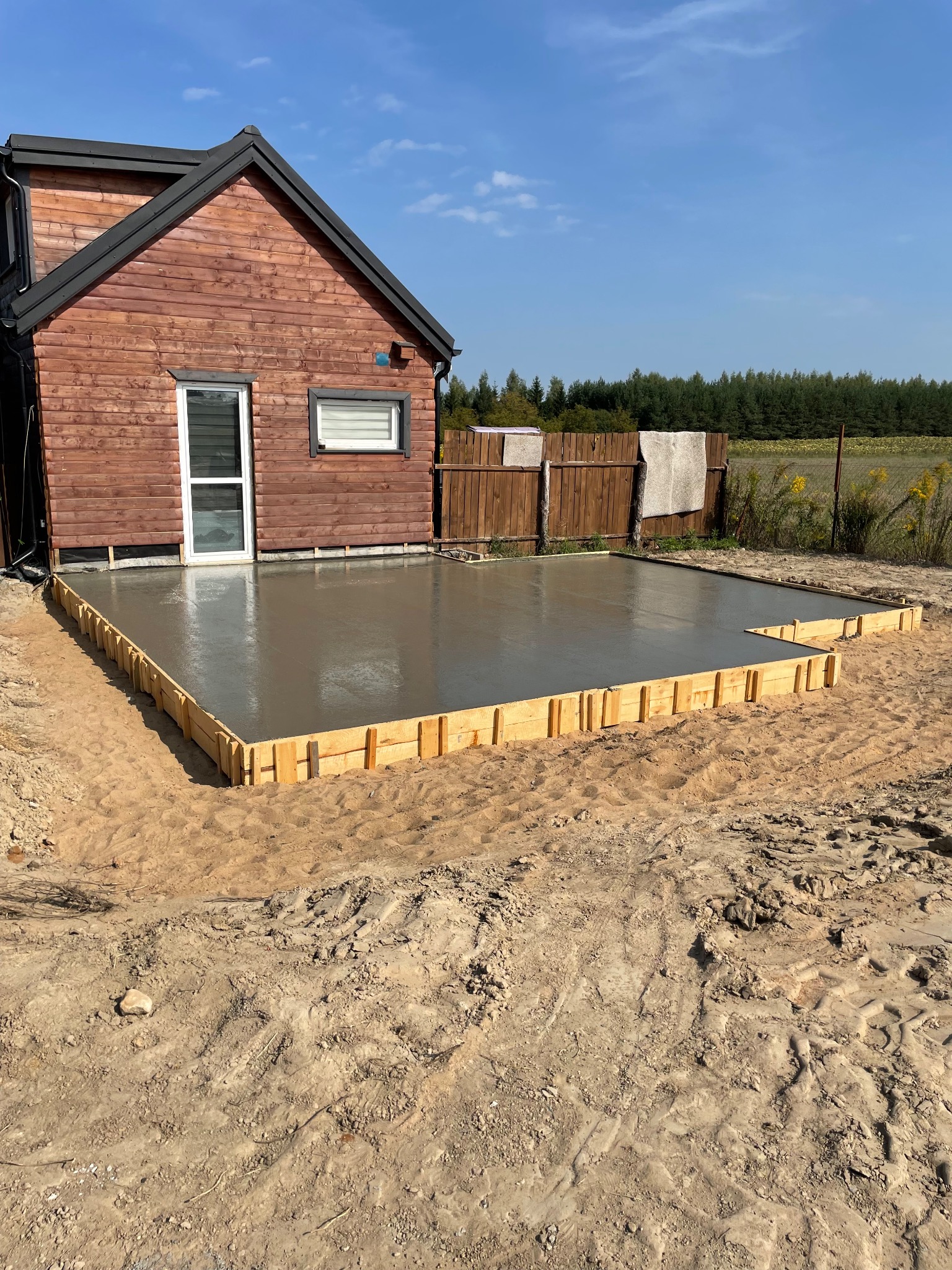 Wylana, świeża płyta fundamentowa z drewnianym szalunkiem przed drewnianym domem z ciemnym dachem w słoneczny dzień.