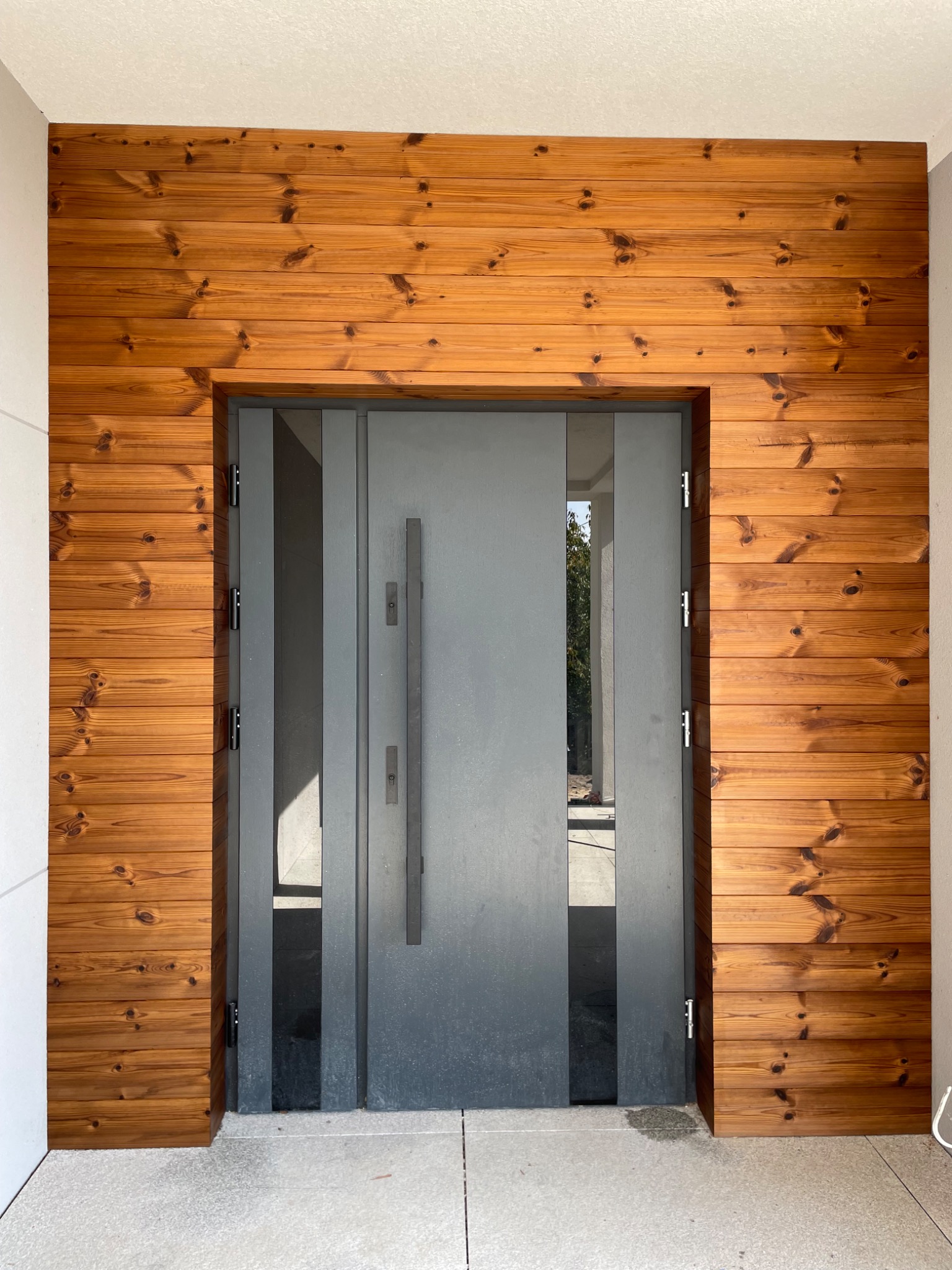 Nowoczesne wejście do domu z szarą, metalową ramą drzwi i okładziną z desek w ciepłym, brązowym odcieniu drewna, tworzącą ramę wokół drzwi.