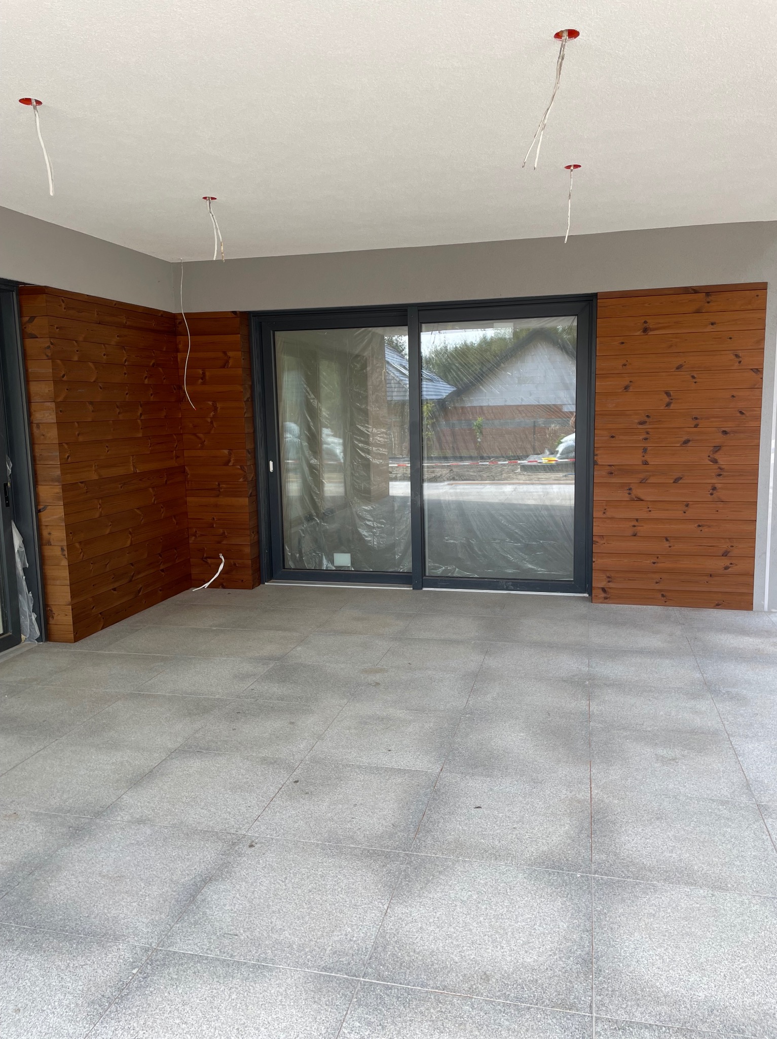 Zadaszony taras z szarą, kwadratową podłogą z płytek, wykończony drewnianą boazerią w odcieniach brązu. Przesuwne drzwi tarasowe zabezpieczone folią ochronną. Widoczne wystające przewody...