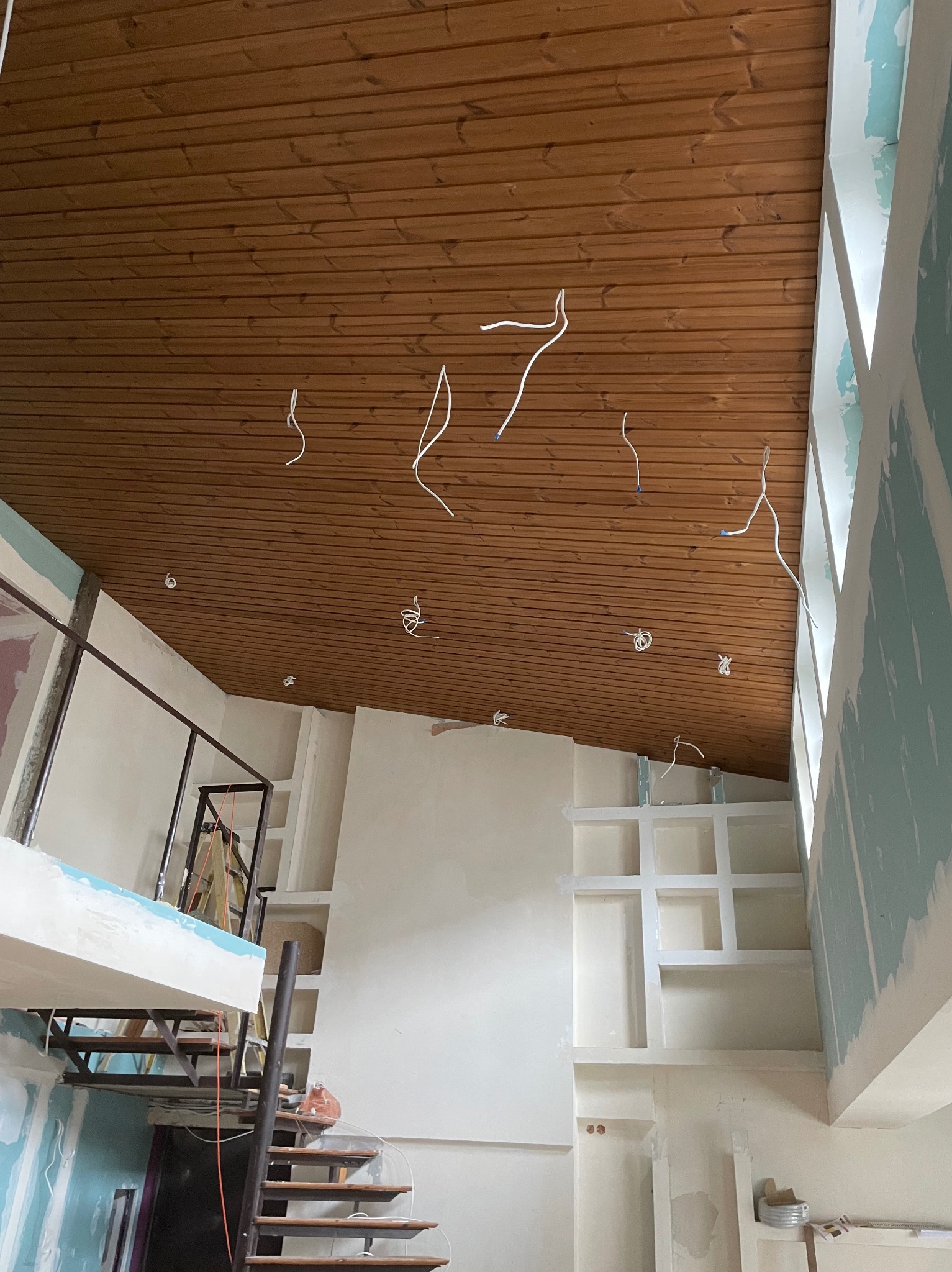 Wnętrze w trakcie remontu: drewniana boazeria na suficie, wystające przewody elektryczne, ściany z płyt kartonowo-gipsowych w trakcie szpachlowania, konstrukcja schodów z metalową balustradą.