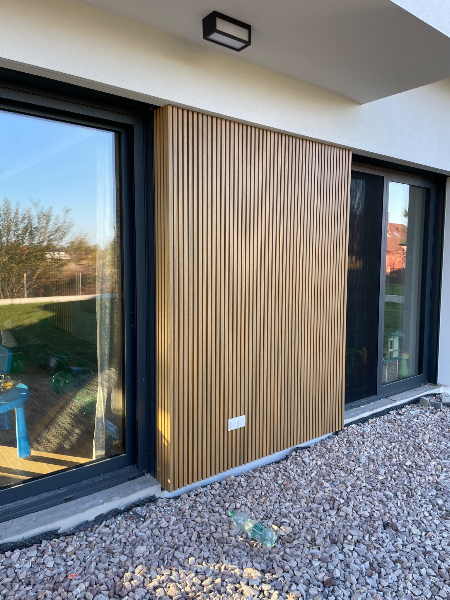 Nowoczesna elewacja domu z drewnianymi lamelami, widoczny fragment okna z czarną ramą i kamienny podkład u podstawy ściany.