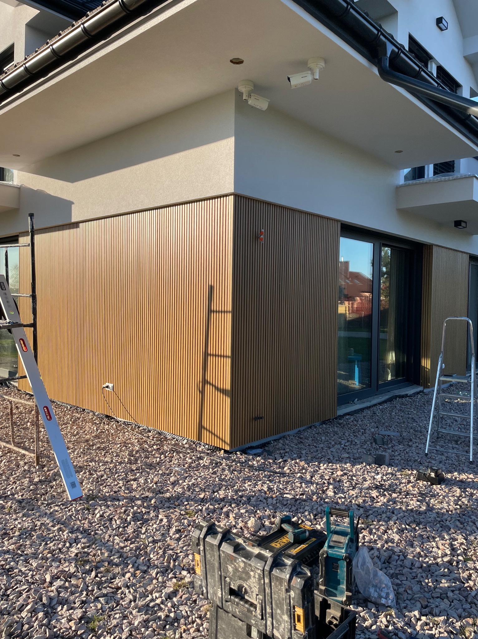 Nowoczesny dom z elewacją z paneli drewnopodobnych, widoczne dwie kamery monitoringu, narzędzia budowlane i drabina oparta o ścianę.