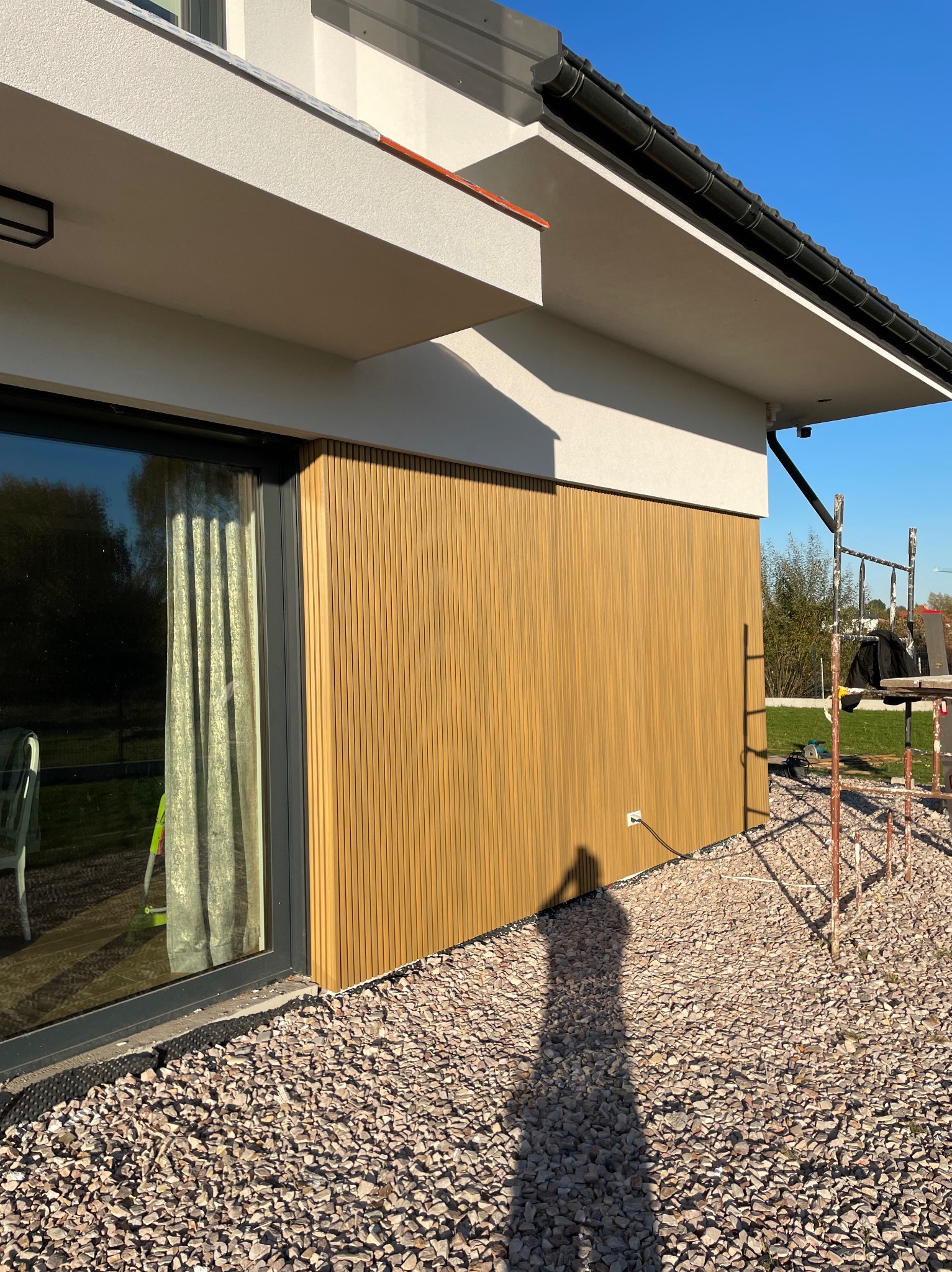 Fragment elewacji domu z pionowymi panelami w odcieniach brązu, widoczny z bliska, z oknem po lewej stronie i rusztowaniem po prawej, na tle błękitnego nieba.