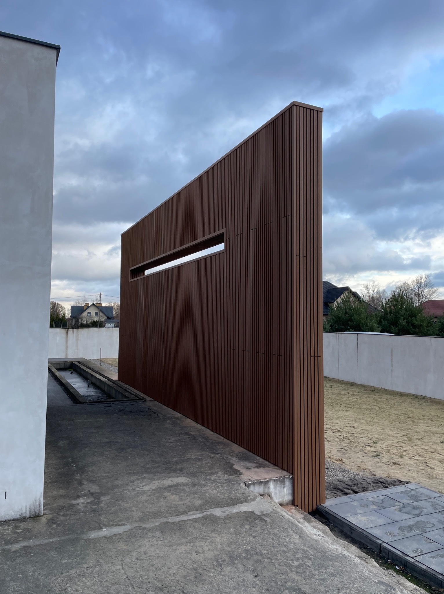 Nowoczesna, pionowa przesłona z brązowych paneli kompozytowych z poziomym oknem, oddzielająca przestrzeń w ogrodzie, z widocznym betonowym podłożem i fragmentem budynku w tle.