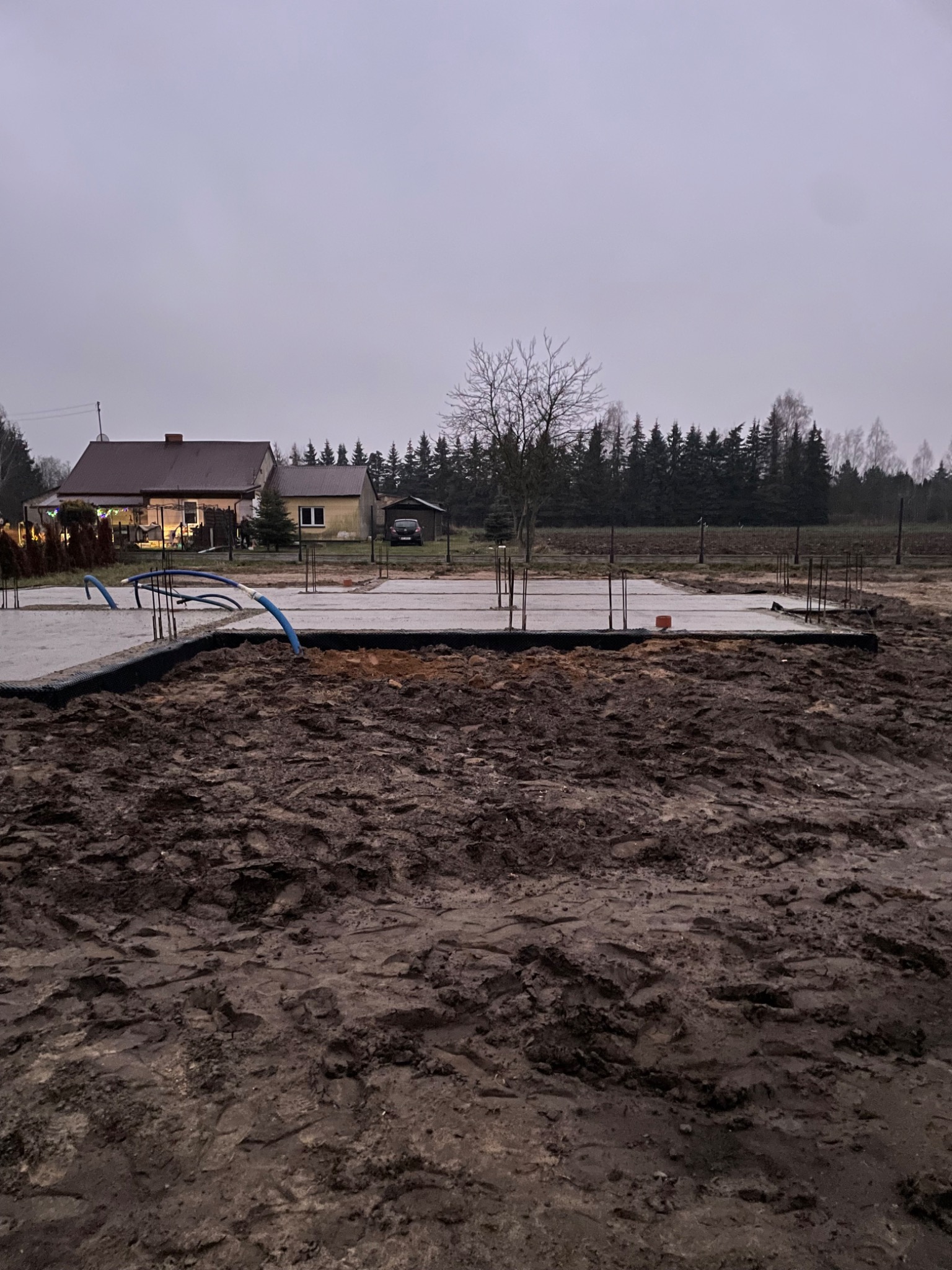 Wylana płyta fundamentowa z wystającymi prętami zbrojeniowymi i niebieskimi rurami instalacji, na pierwszym planie błotniste podłoże, w tle zabudowania jednorodzinne i las w pochmurny dzień.