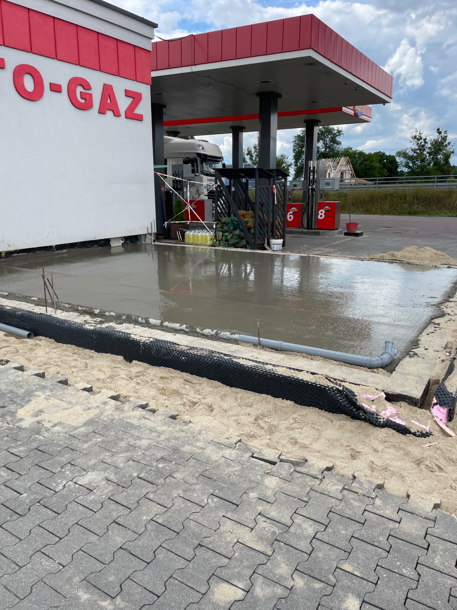 Wylewka betonowa przy stacji benzynowej, widoczne kostki brukowe, rury odpływowe i elementy izolacyjne. Etap prac budowlanych na zewnątrz budynku.