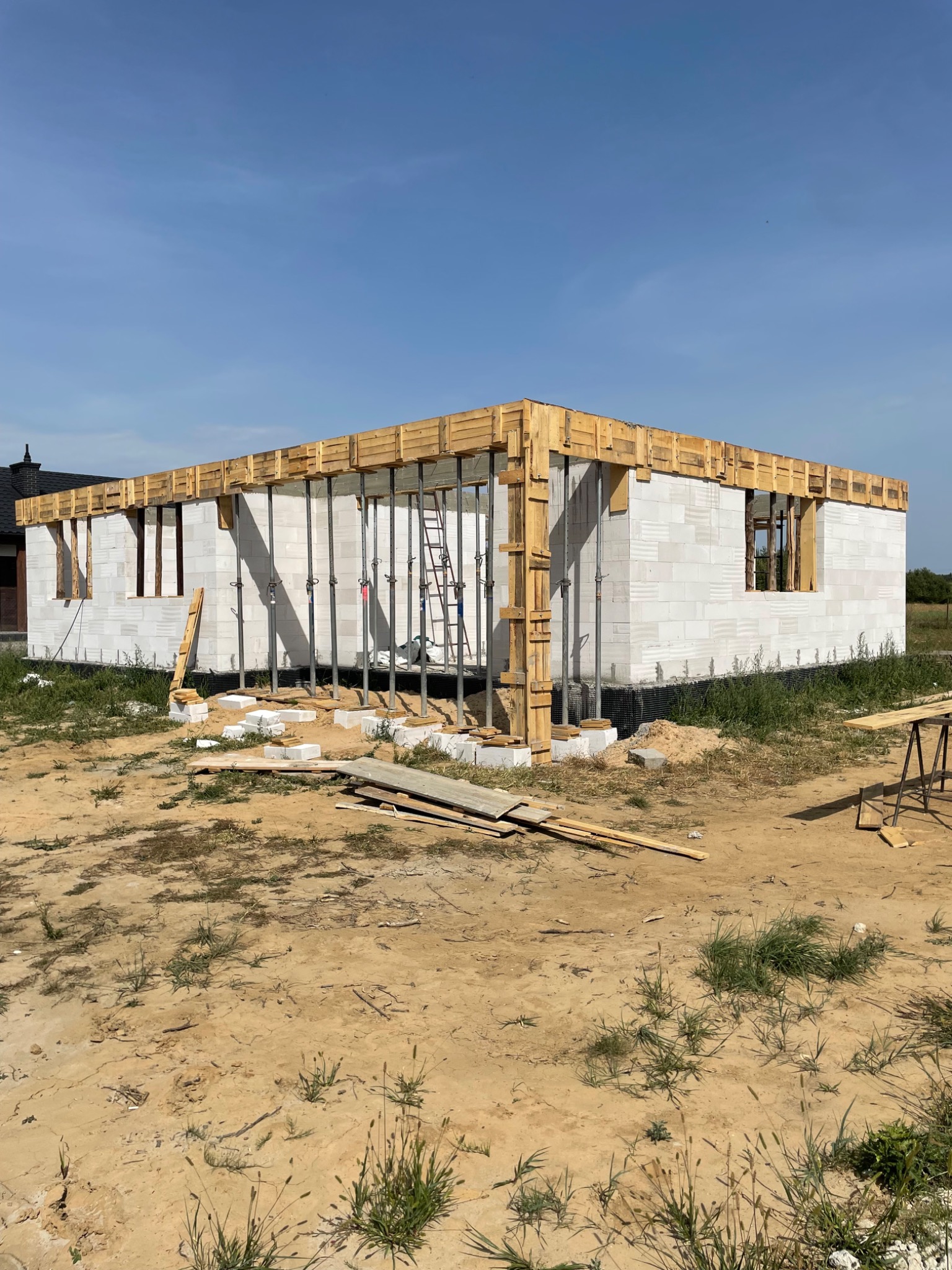 Wznoszenie ścian z białych bloczków betonu komórkowego. Widoczne drewniane szalunki i podpory stropu. Częściowo wykończony budynek w stanie surowym otwartym.