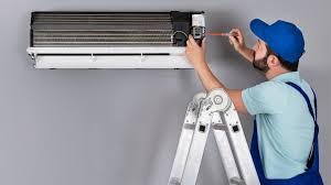 Mężczyzna w niebieskiej czapce serwisuje klimatyzator ścienny, używając śrubokręta. Stoi na aluminiowej drabinie rozstawnej na tle szarej ściany.