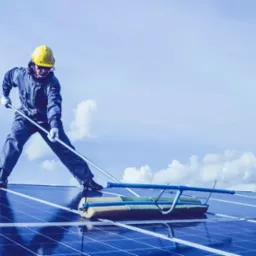 Pracownik w kombinezonie i żółtym kasku czyści panele słoneczne na dachu za pomocą szczotki na długim kiju na tle błękitnego nieba z chmurami.