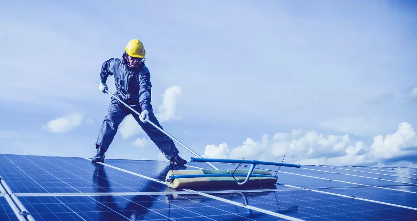 Pracownik w kombinezonie i żółtym kasku czyści panele słoneczne na dachu za pomocą szczotki na długim kiju na tle błękitnego nieba z chmurami.