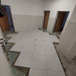 Patryk Frątczak usługi remontowo wykończeniowe - Układanie jasnych, kwadratowych płytek podłogowych w pomieszczeniu z trzema parami drzwi; widoczne krzyżyki dystansowe i fragment instalacji wodnej przy ścianie.