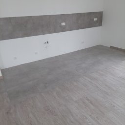Patryk Frątczak usługi remontowo wykończeniowe - Wykończone wnętrze: szare płytki na ścianie i podłodze przechodzące w panele podłogowe o jasnym, drewnopodobnym wzorze. Widoczne gniazdka elektryczne i przyłącza wodne.