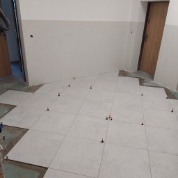 Patryk Frątczak usługi remontowo wykończeniowe - Układanie jasnych, kwadratowych płytek podłogowych z użyciem poziomujących klipsów i klinów w pomieszczeniu z dwukolorowymi ścianami i drewnianymi drzwiami.