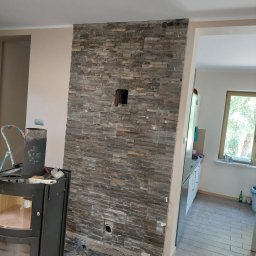 Patryk Frątczak usługi remontowo wykończeniowe - Ściana wyłożona kamieniem dekoracyjnym w trakcie montażu, widoczne otwory na instalacje, fragment kominka i zabezpieczona folią podłoga.