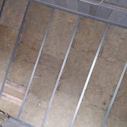 Patryk Frątczak usługi remontowo wykończeniowe - Montaż izolacji z wełny mineralnej w metalowym stelażu na ścianie, widoczne fragmenty surowego sufitu i podłogi.