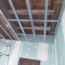 Patryk Frątczak usługi remontowo wykończeniowe - Montaż stelażu sufitu podwieszanego z płyt gipsowo-kartonowych, widoczne drewniane belki stropu i częściowo położone płyty, drabina przystawiona do ściany.