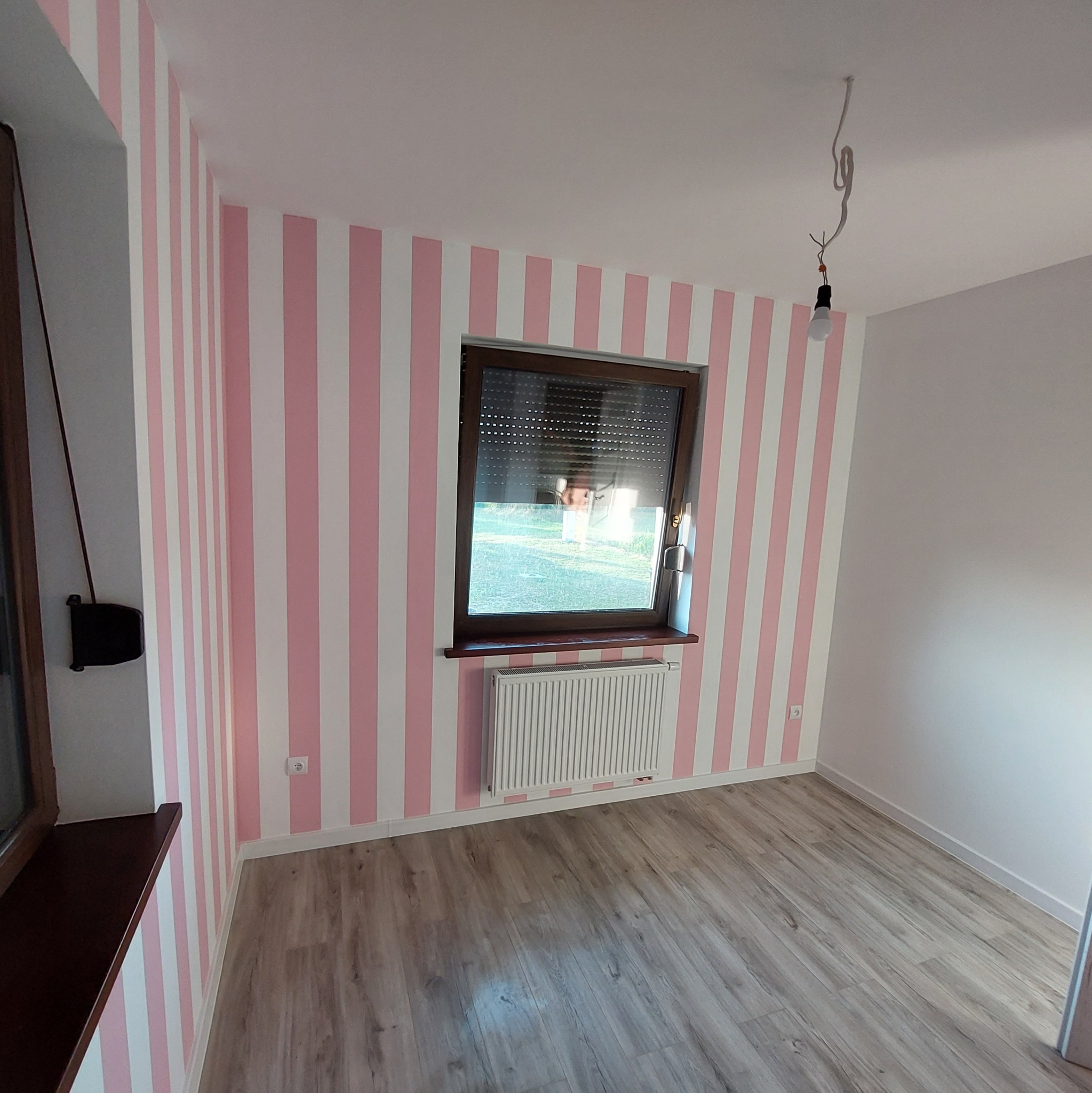 Pokój po malowaniu ścian w różowo-białe pasy, z oknem z roletą, grzejnikiem i panelami na podłodze, widoczna wisząca żarówka bez oprawy.
