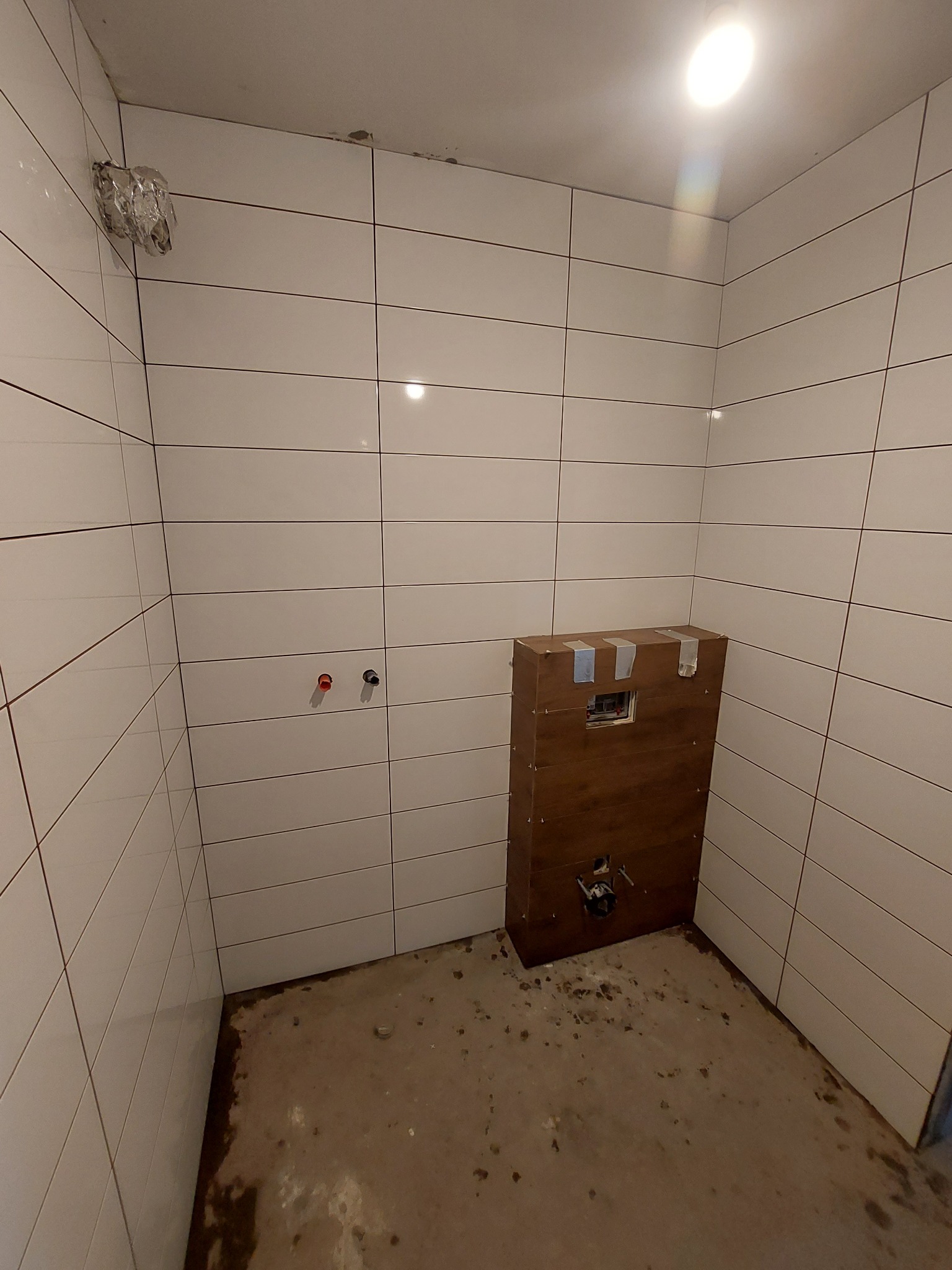 Wnętrze łazienki w trakcie remontu: ułożone białe, poziome płytki, przygotowane przyłącza wodne, zabudowa stelaża WC z płyt OSB zabezpieczona taśmą, surowa podłoga przed wylaniem wylewki.