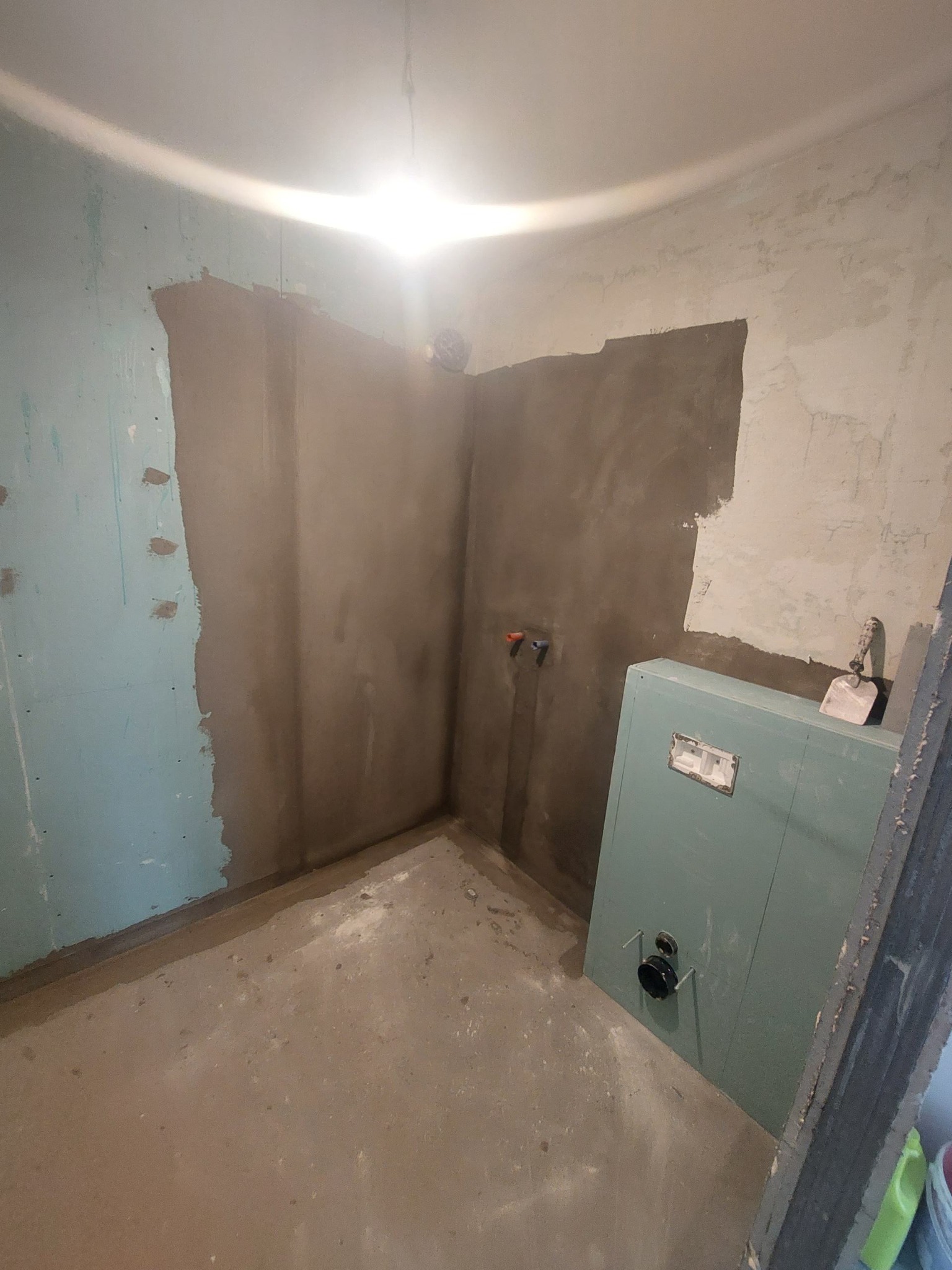 Surowe wnętrze łazienki w trakcie prac wykończeniowych; częściowo otynkowane ściany, widoczne instalacje, zabudowa stelaża podtynkowego i narzędzia.