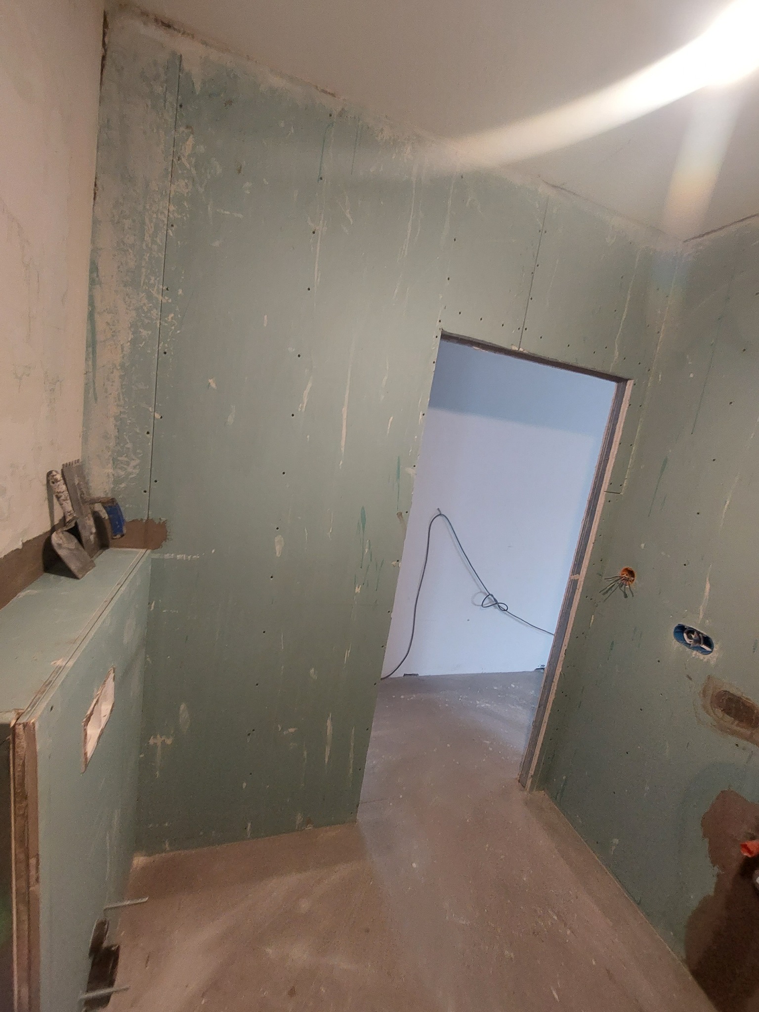 Wnętrze w trakcie prac przygotowawczych: ściany z płyt gipsowo-kartonowych, widoczne narzędzia szpachlarskie na półce, instalacja elektryczna w puszce, surowa podłoga.