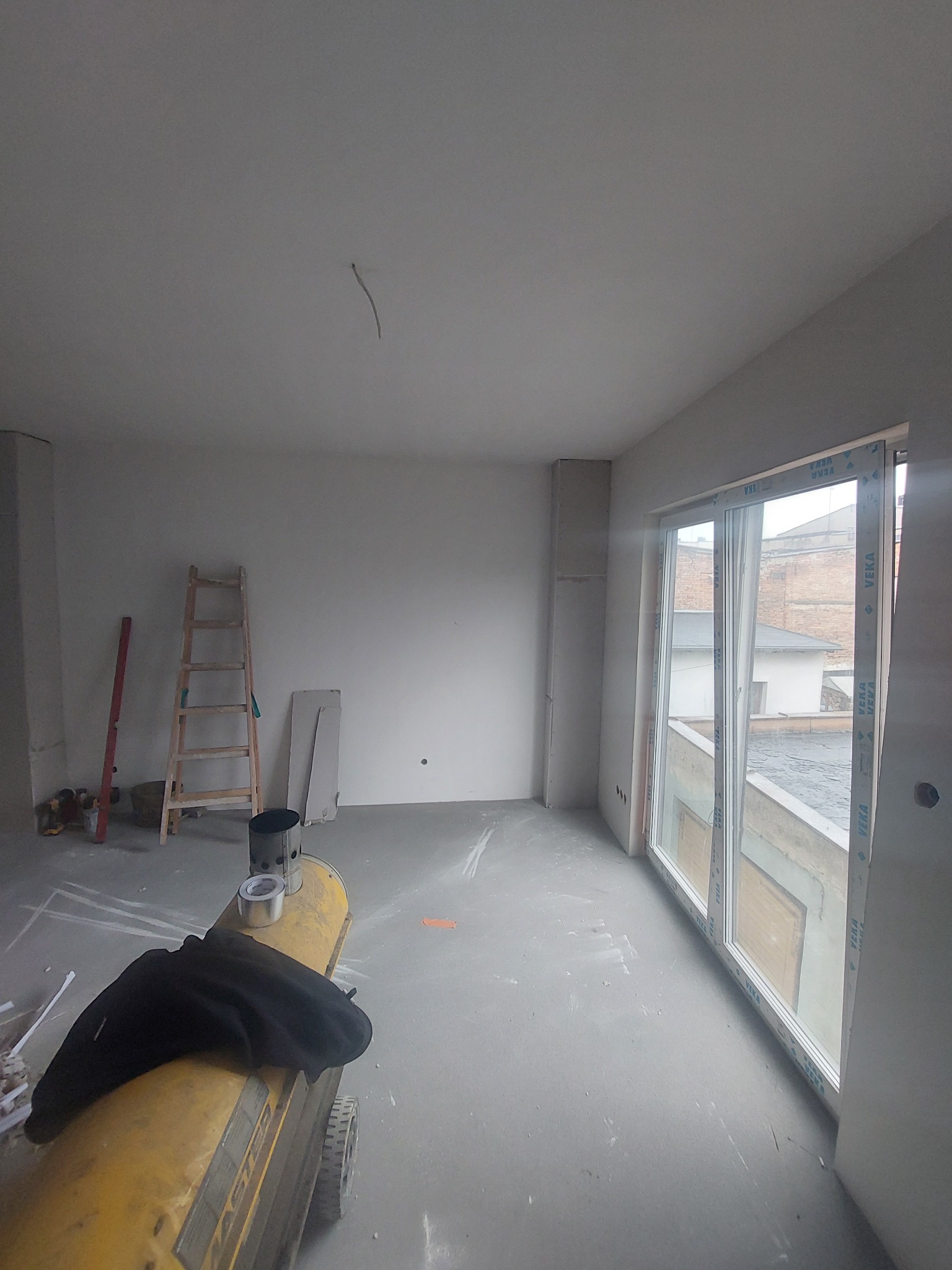 Wnętrze pokoju w trakcie remontu, ściany przygotowane pod malowanie lub gładzie, widoczna drabina, poziomica, grzejnik budowlany i okno balkonowe.