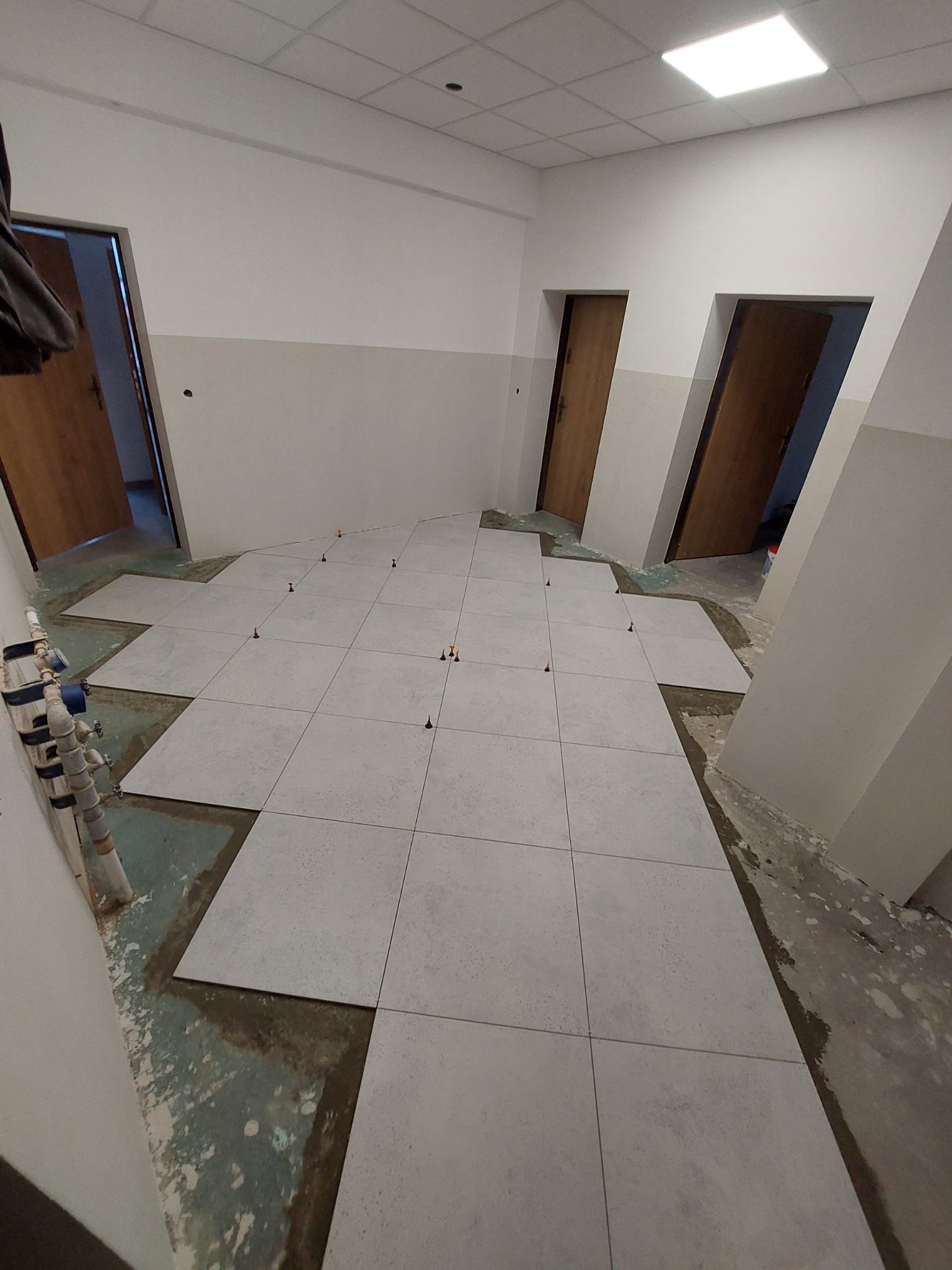 Układanie jasnych, kwadratowych płytek podłogowych w pomieszczeniu z trzema parami drzwi; widoczne krzyżyki dystansowe i fragment instalacji wodnej przy ścianie.
